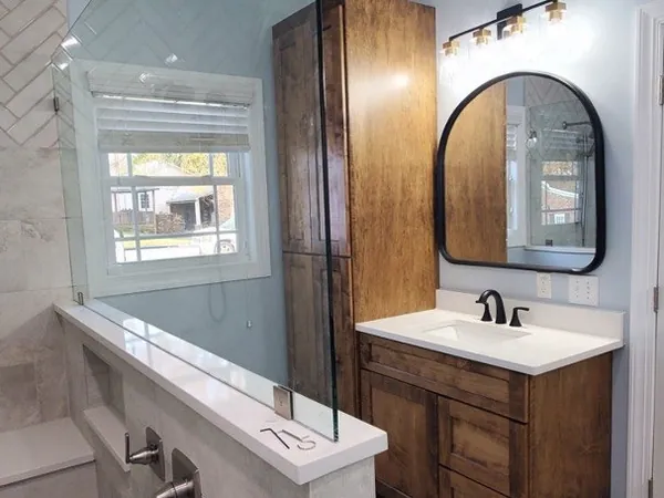 A finished bathroom: a walk-in tile shower with a glass panel on a tiled half-wall, and a wood vanity with a round mirror across the room.