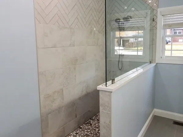 A finished walk-in tile shower with a herringbone tile back wall, a glass panel set on a tiled half-wall, and a river-pebble floor behind a low tiled curb.