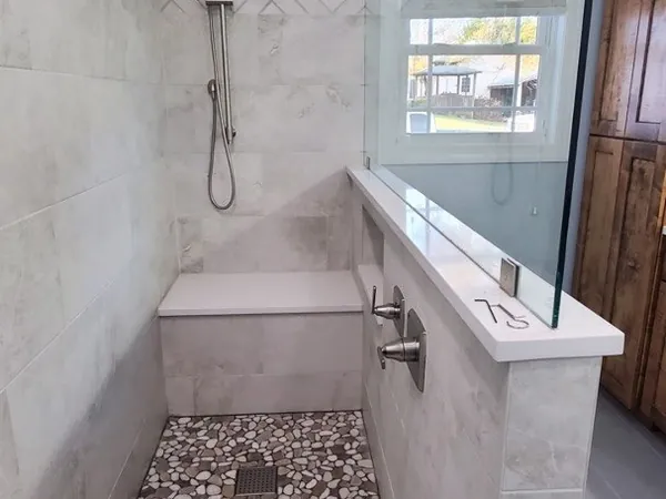 Inside a finished tile shower: a herringbone tile wall, a tiled bench, a hand shower on a slide bar, and a river-pebble floor falling to a square drain.